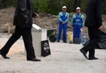 Two construction workers wearing hard hats looking as two men dressed in dark suits pass by.