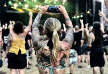 A group of people turned away from the camera to cface a stage with their arms raised in the air, A woman in the foreground has tatoos on her back and arms and is taking pictures with her mobile.