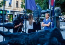 A person interviewed by a journalist in front of a camera in a city square with a section of a sculpture and flags in the foreground.